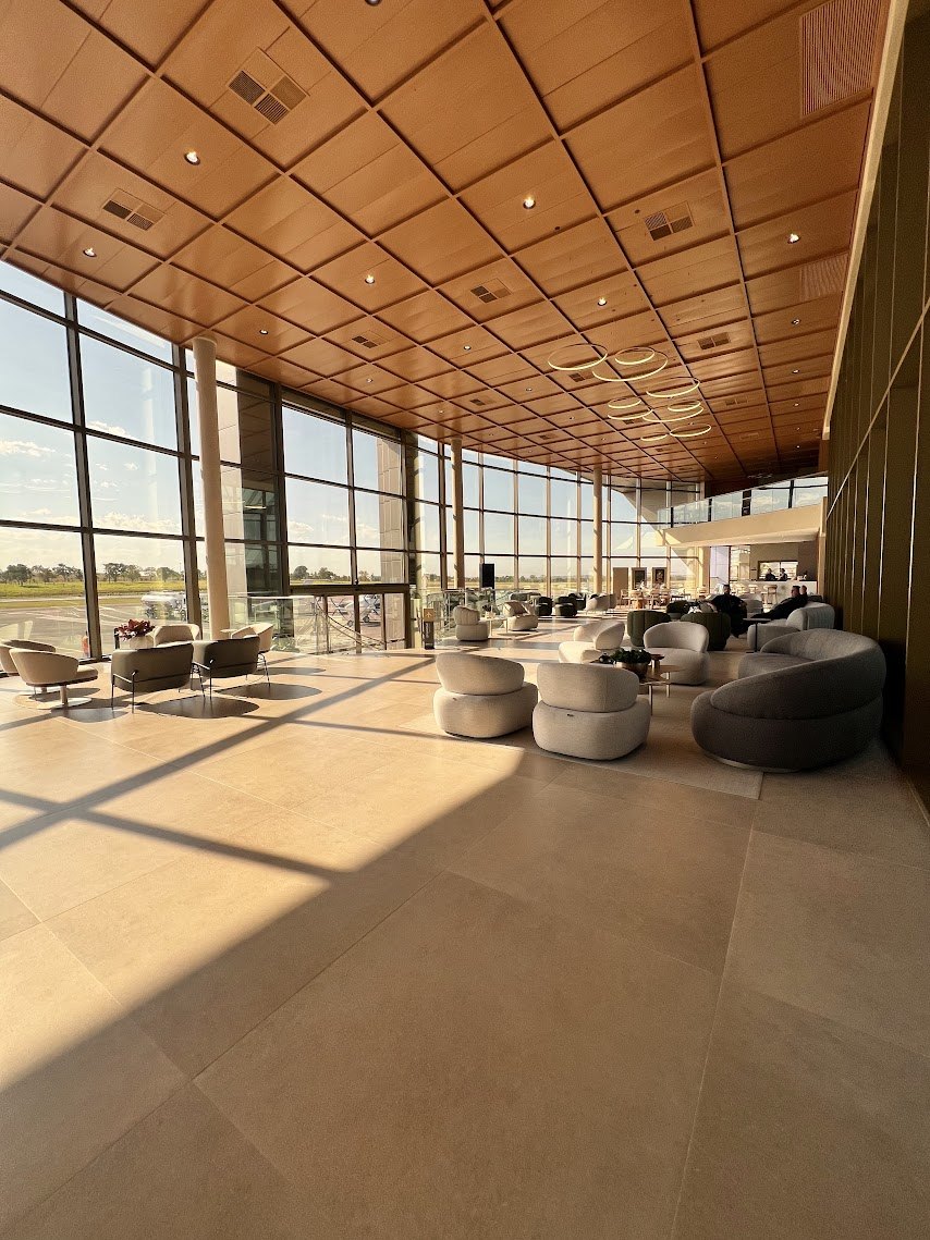 Sala de espera do terminal, com teto de madeira, poltronas claras e fachada de vidro voltada para a pista