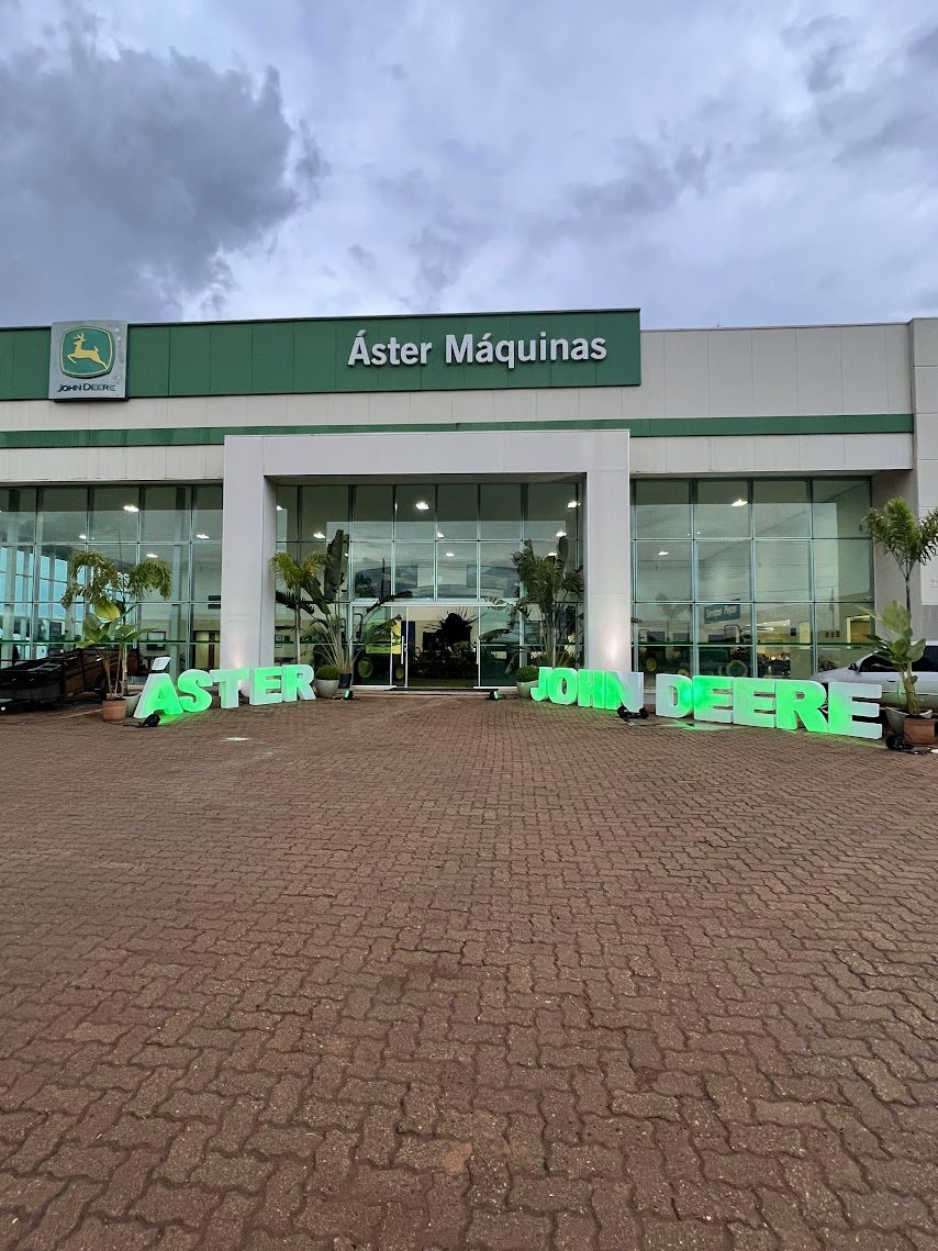 Fachada da concessionária Áster Máquinas John Deere, com o letreiro da marca no pátio, sob céu carregado