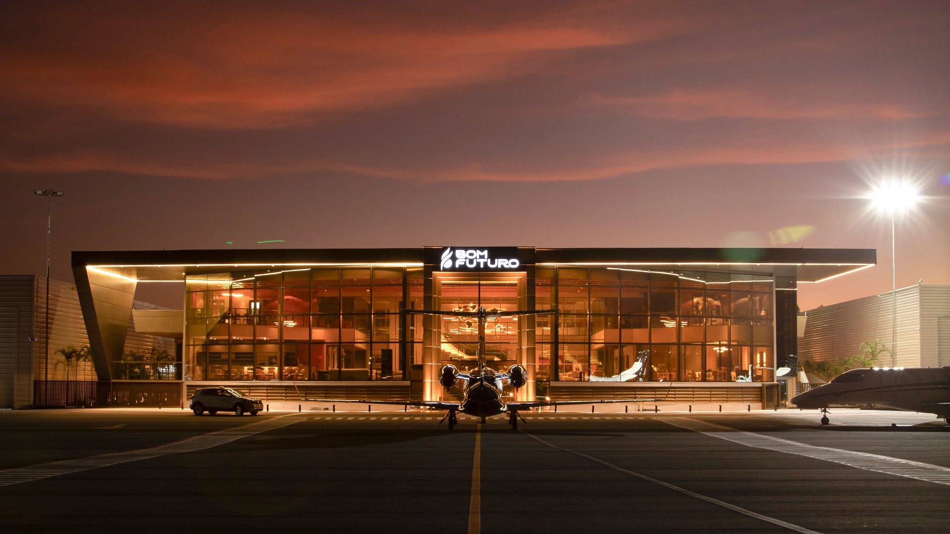 Terminal executivo Bom Futuro iluminado ao anoitecer, com um jato estacionado de frente para a fachada de vidro