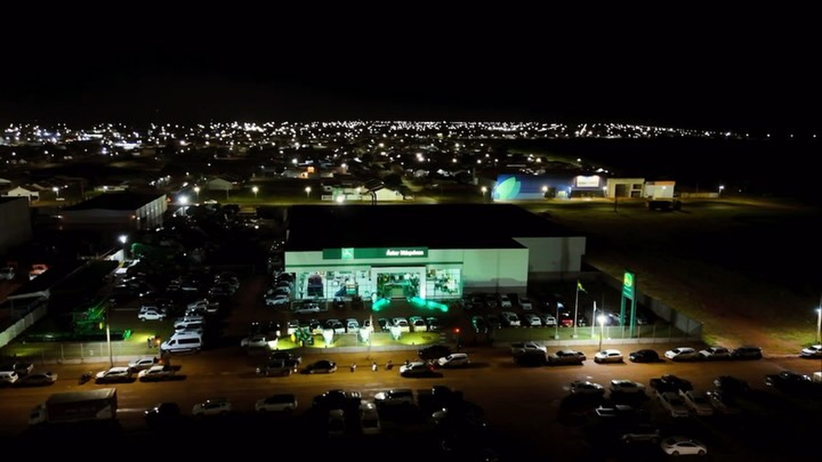 Vista aérea noturna da concessionária Áster John Deere iluminada, com o pátio cheio de carros